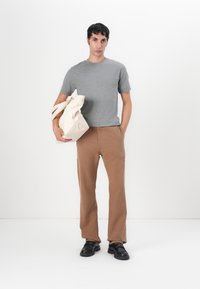 Grijze T-shirt met korte mouwen, bruine rechte broek en zwarte schoenen. Houdt een lichtbeige tote bag vast met korte handvatten.