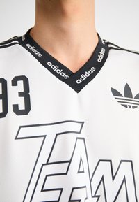 Balta sportinė marškinė su juodais akcentais, turi V formos iškirptę, ryškų "TEAM" spaudą ir "adidas" logotipą. Textūruota medžiaga, kuri atrodo kvėpuojanti.