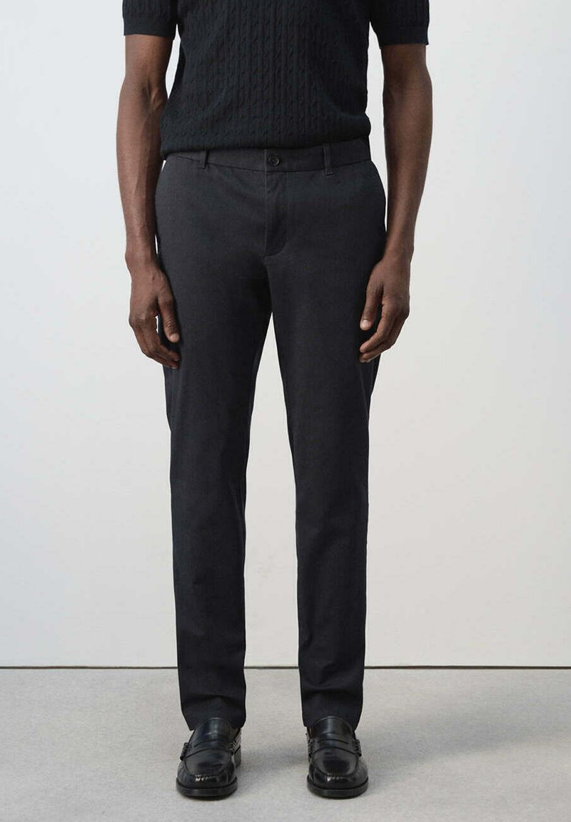 Homme portant un pantalon noir ajusté, un haut court noir tressé à manches courtes et des mocassins en cuir noir, debout devant un fond uni.