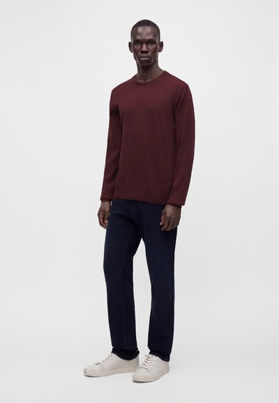Homme debout portant un pull bordeaux à manches longues, un pantalon foncé et des baskets blanches sur un fond clair uni.