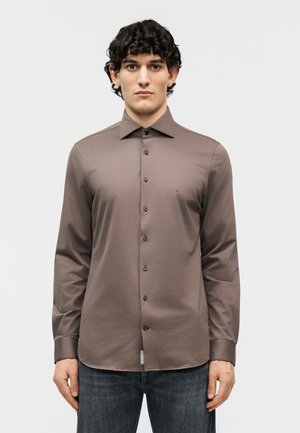 Νεαρός άνδρας με σγουρά μαλλιά φορά καφέ πουκάμισο με κουμπιά και σκοτεινό τζιν, στέκεται όρθιος μπροστά σε απλό ανοιχτό φόντο.