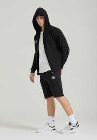 Felpa nera con zip e cappuccio, abbinata a pantaloni corti sportivi neri. Accenti del logo bianco su entrambi i capi. Il modello indossa sneakers e calze bianche.