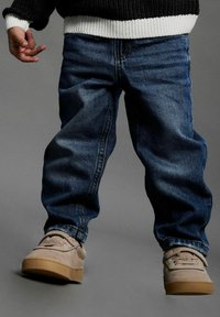 Plave traperice slobodnog kroja, u kombinaciji s bež antilop cipelama s teksturiranim završetkom i gumenim potplatima. Detalj tamnog pletenog džempera vidljiv.