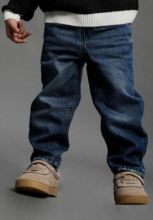 Zila džinsa bikses ar vaļīgu piegriezumu, apvienotas ar brūnā zamša apaviem ar tekstūru un gumijas zole. Redzams tumšs adīts džemperis.