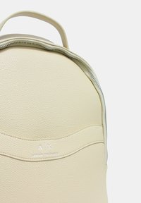 Zaino in ecopelle color crema con superficie testurizzata, parte superiore curva, chiusura con zip e un logo goffrato discreto sulla parte anteriore.