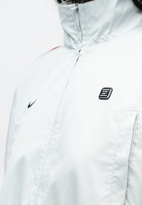 Valge kerge jope kõrge kraega, ees tõmblukk ja kontrastsed punased detailid. Omab Nike musta logo ja väikest tekstureeritud plaastrit.