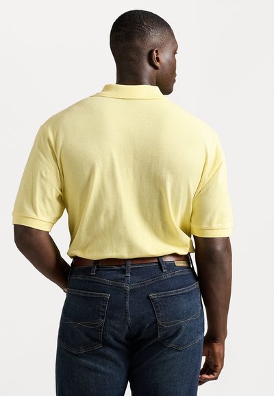 Camisa polo amarela de mangas curtas, com tecido texturizado e gola clássica, combinada com jeans de ganga azul-escura com costuras em contraste.