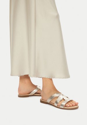 Sandalias planas doradas metálicas con tres tiras anchas, un detalle floral y suela plana, combinadas con un vestido beige fluido.