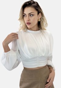 Weißes, gekürztes Oberteil mit Puffärmeln, plissiertem Design und geraffter Textur an der Taille. Getragen mit einem hellbraunen, hoch taillierten Rock.
