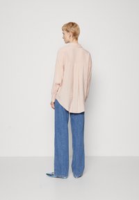 Camicia rosa chiaro a maniche lunghe con colletto a bottoni, vestibilità rilassata e orlo curvo, abbinata a jeans blu in denim a gamba larga e scarpe blu.