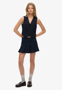 Granatowa kamizelka w prążki z dekoltem w serek i zapięciem na guziki, w połączeniu z dopasowaną plisowaną mini spódnicą. Stylizowana z szarymi skarpetami w paski i loafersami.
