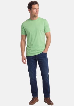 Uomo con T-shirt verde chiaro a maniche corte, jeans blu scuro e sneakers marroni in pelle, in piedi con una mano in tasca.