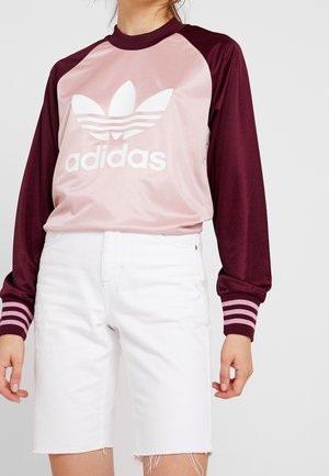Femme portant un sweat-shirt Adidas rose et bordeaux avec logo blanc et un short en jean blanc jusqu'au genou sur un fond uni.