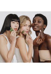 Trois femmes diverses utilisent des rouleaux pour le visage et des outils gua sha sur leur visage, portant des vêtements minimalistes aux tons neutres sur un fond uni.