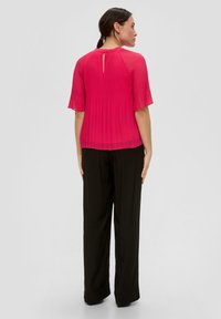 Geverde, felroze top met een sleutelgat aan de achterkant, gecombineerd met losse zwarte broek, wat een vloeiende silhouet laat zien.