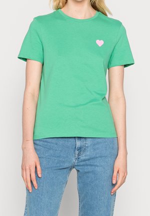 Zielony bawełniany t-shirt z krótkimi rękawami, okrągłym dekoltem i małą różową grafiką serca na lewej piersi. Noszony z jasnoniebieskimi dżinsami.