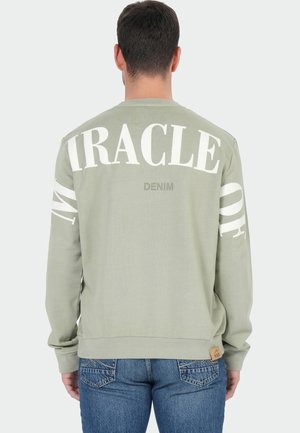 Mann trägt salbegrünen Pullover mit den Aufdrucken "MIRACLE" und "DENIM" auf dem Rücken, kombiniert mit blauen Jeans, von hinten gezeigt.