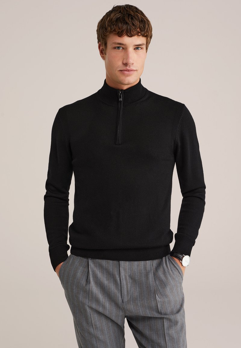 Schwarzer Reißverschluss-Pullover aus Strickstoff, mit hohem Kragen und gerippten Bündchen, kombiniert mit hellgrauen gestreiften Hosen.