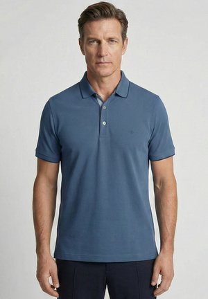 Mann mit kurzem braunen Haar, der ein eng anliegendes blaues Poloshirt und dunkle Hosen trägt, steht vor einem schlichten hellen Hintergrund.