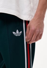 Main gauche reposant à côté d'un pantalon de survêtement Adidas noir avec des bandes latérales blanches, rouges et noires et un logo trefoil blanc, associé à une chemise blanche.