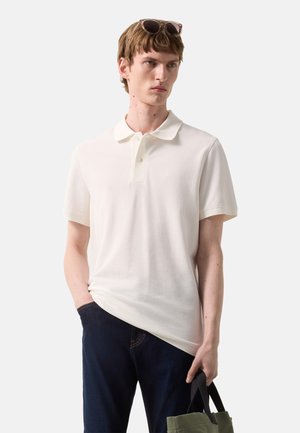 Jonge man met wit poloshirt en donkere jeans, draagt een groene tas, met een zonnebril op zijn hoofd.