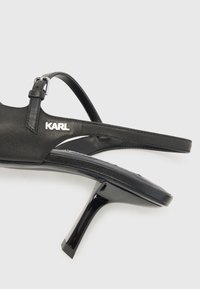 Mustat nahkaiset korokepohjasandaalit, joissa on hoikat korkeakengät, säädettävä nilkkaremmari ja hopeinen "KARL" logo sivulla. Sujuva rakenne ja minimalistinen muotoilu.