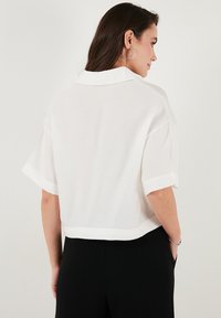 White cropped shirt with short sleeves, collared neckline, and a smooth texture. Features rolled sleeve cuffs and a clean, minimalist design.