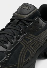 Detalhe de um sapato de corrida ASICS preto e oliva escuro, apresentando tecido em malha, sobreposições sintéticas e atacadores pretos amarrados em padrão cruzado.