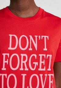 Gros plan d'une personne portant une chemise rouge avec un grand texte rose indiquant "N'OUBLIEZ PAS D'AIMER."