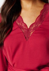 Vêtement rouge avec un décolleté en V profond orné de dentelle. Tissu doux avec une texture lisse. Comprend une ceinture en tissu à la taille.