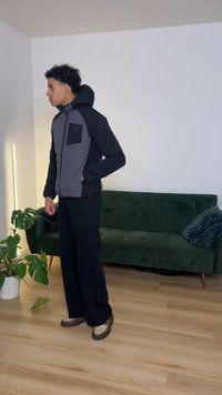 Giacca imbottita nera e grigia con zip frontale, cappuccio e taschino sul petto. Abbinata a pantaloni neri larghi e scarpe marroni.