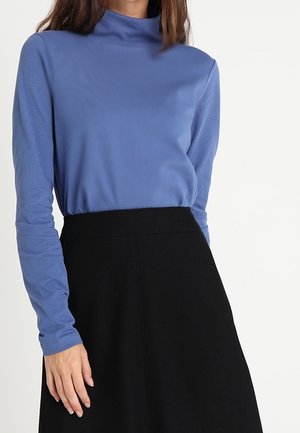 Mujer que lleva un suéter azul de manga larga con cuello alto, metido dentro de una falda negra de línea A de talle alto, sobre un fondo sencillo.