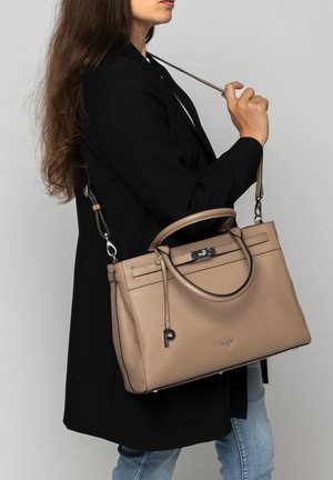 Woman in black coat holding beige leather handbag with shoulder strap and top handles, standing against a plain light background.