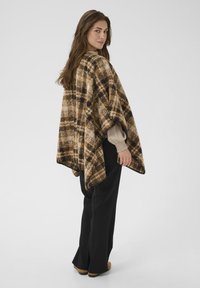 Rutig brun poncho med ett mjukt, texturerat tyg och en kontrasterande svart kant, bärs över en ljus blus och matchas med svarta byxor.