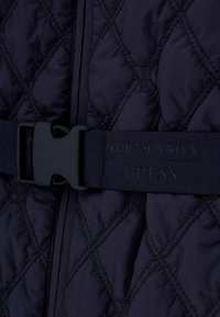 Gros plan sur un tissu matelassé foncé avec des surpiqûres en forme de losange et une ceinture marine dotée d'une boucle en plastique noire et du marquage "North Sails GUESS".