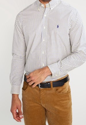 Mężczyzna w białej i niebieskiej koszuli w kratę, wpuszczonej w brązowe spodnie sztruksowe z czarnym paskiem, ręka poprawiająca klamrę paska.