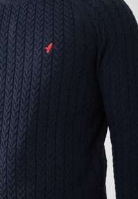 Marineblauer Pullover mit strukturiertem Kabelstrickmuster. Gibt es mit einem kleinen roten Vogel-Emblem auf der Brust, das einen Kontrast zum dunklen Stoff bietet.