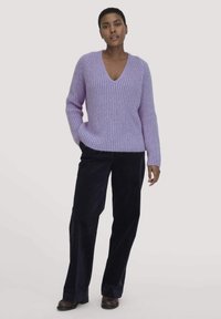 Lila gerippter Strickpullover mit V-Ausschnitt, kombiniert mit dunklen, weit geschnittenen Hosen. Das Material wirkt weich und strukturiert, was das Gesamt-Design unterstreicht.