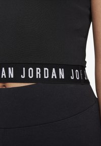 Czarny krótki top o gładkiej fakturze i szerokim elastycznym pasku z napisem "JORDAN" w kolorze białym. Krój jest przylegający i sportowy.