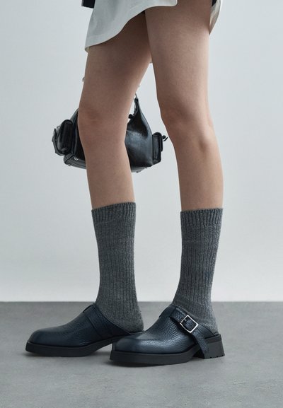 Zapatos de cuero azul con un acabado texturizado, una hebilla lateral y una suela plana ancha, combinados con calcetines grises de canalé.