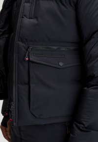 Czarna pikowana kurtka z dużą kieszenią na zamek błyskawiczny i zatrzaski, oznaczoną napisem "Zavetti Canada", z suwakami w kształcie liścia klonu na rękawie i z przodu.