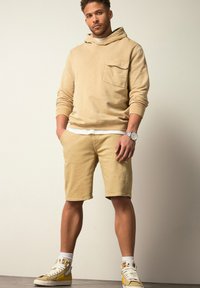 Beige hoodie met een voorzak, gecombineerd met beige shorts. Witte onderkast zichtbaar. Schoeisel is crème sneakers met gele accenten.