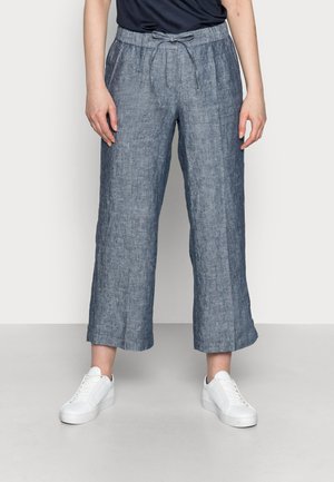 Osoba ubrana w luźne, sięgające kostek szare lniane spodnie z wiązanym pasem i białe trampki, stojąca na tle jednolitego tła.