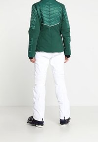 Veste de ski matelassée verte au design structuré, associée à un pantalon de ski blanc avec des accents texturés. Bottes de ski noires visibles à la base.