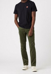 Černé tričko s krátkým rukávem a malým červeným logem, spárované s olivově zelenými kordovými kalhotami a bílými teniskami.