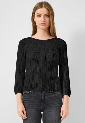 Vrouw met steil haar, gekleed in een zwarte trui met lange mouwen en een donkere grijze spijkerbroek, staand tegen een effen lichte achtergrond.