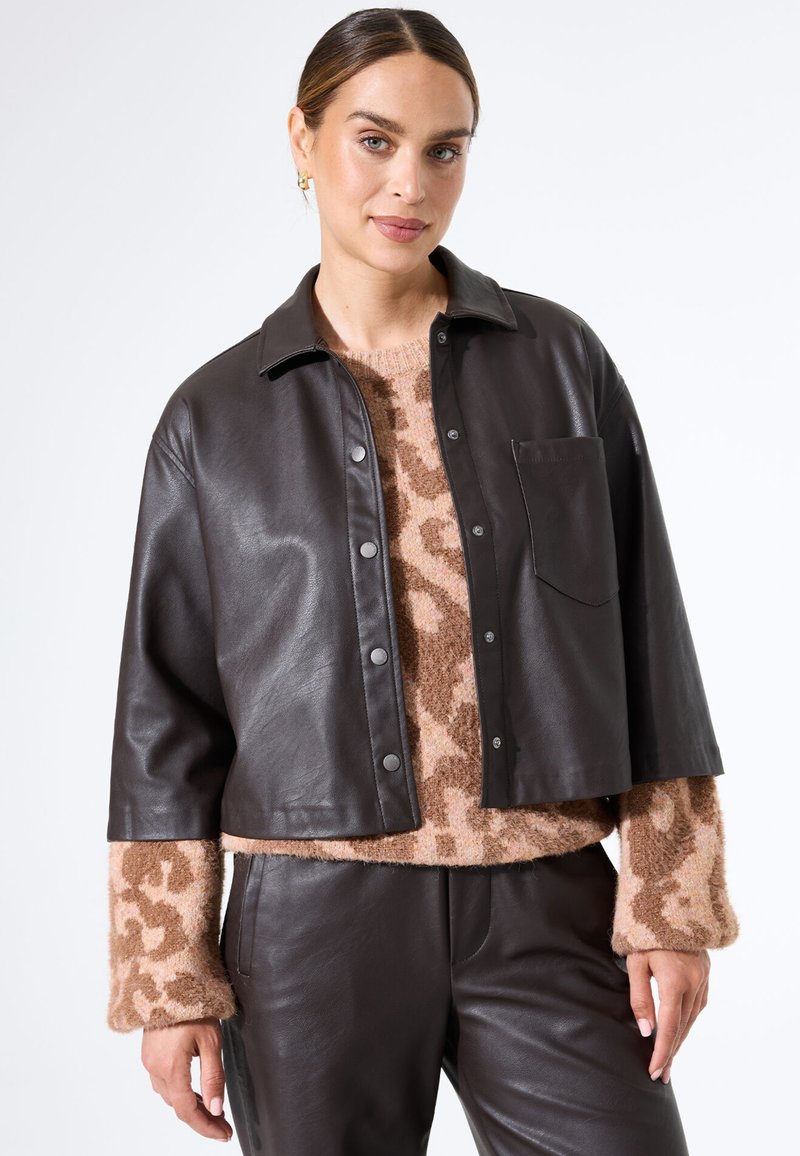 Bruine leren jas met een cropped ontwerp, drukknoopsluiting en een enkele borstzak, gelaagd over een trui met luipaardpatroon.