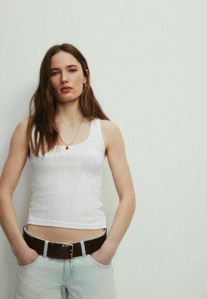 Ung kvinde med langt brunt hår iført en hvid ærmeløs top, lyse jeans og et mørkt bælte, står mod en ensfarvet lys væg.