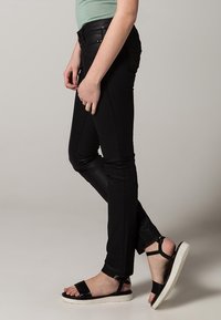 Vaqueros negros ajustados con una textura suave y un brillo sutil, combinados con sandalias negras que tienen una suela blanca y un diseño de tiras.