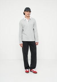 Grijze longsleeve polo met witte kraag, gecombineerd met zwarte jeans en rood-witte sneakers. Model draagt een donkere pet. Eenvoudige achtergrond.